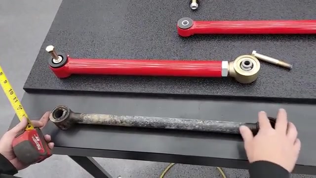 New red Core 4x4 Camp Series control arm with Johnny Joint next to the worn factory 4Runner arm on the workbench with a tape measure showing the 21-inch center-on-center measurement