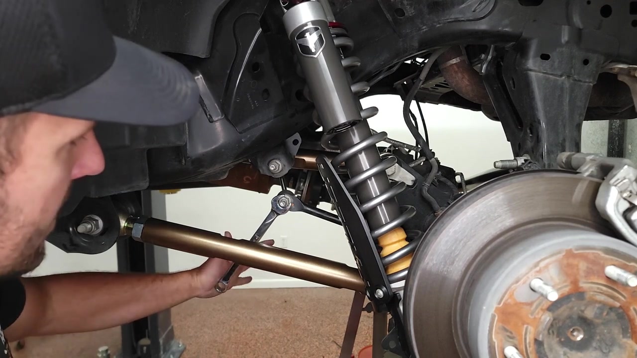 Spence underneath the 2022 Ford Bronco working on the rear suspension, showing the factory sway bar end link connected near the coilover shock