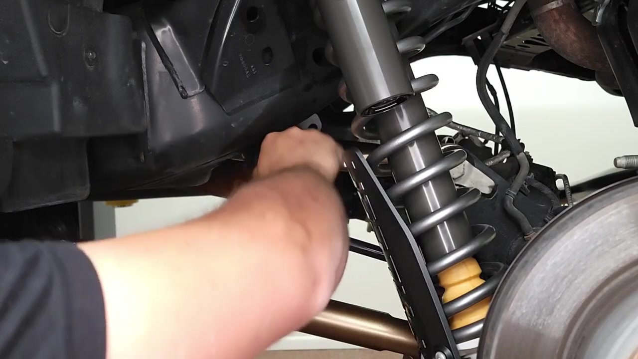 Close-up of hands removing the factory rear sway bar end link on the Bronco, showing the coilover spring and suspension components