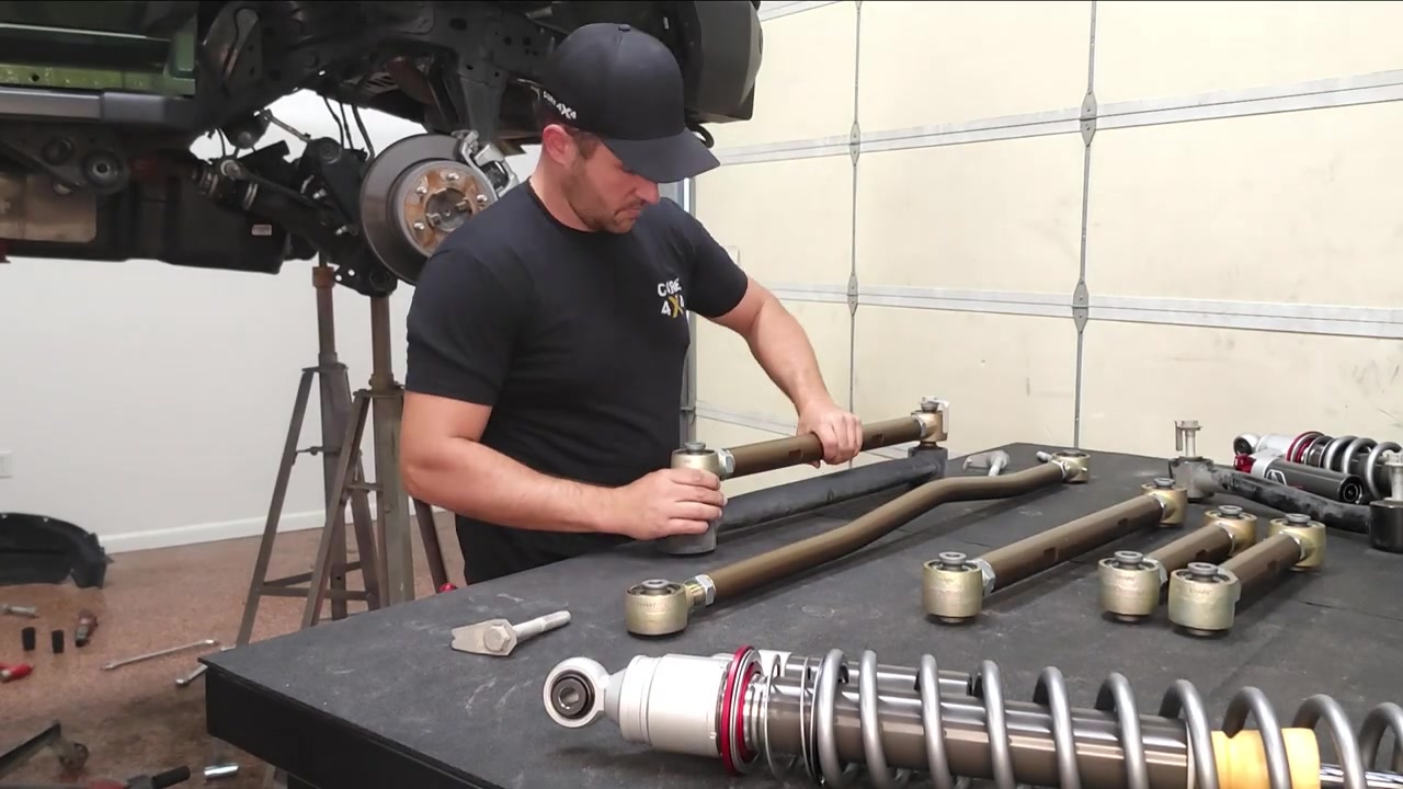Spence adjusting rear control arm length on the workbench, matching left and right arms side by side with bronze Core 4x4 arms and Falcon coilovers visible