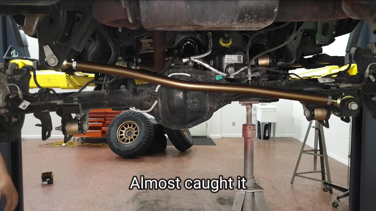 Rear view under the Ford Bronco showing the new bronze track bar installed across the rear axle with Falcon coilover visible and Method bronze wheel on the ground