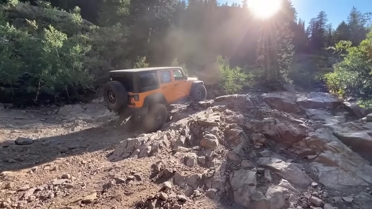 Orange Jeep Wrangler crawling over rocks on a mountain trail with sun flare through the trees