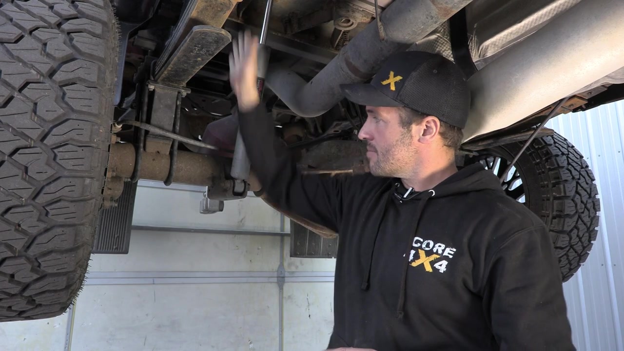 Spence under the Ram 3500 pointing out the rear axle area and measuring bracket placement with a plumb bob