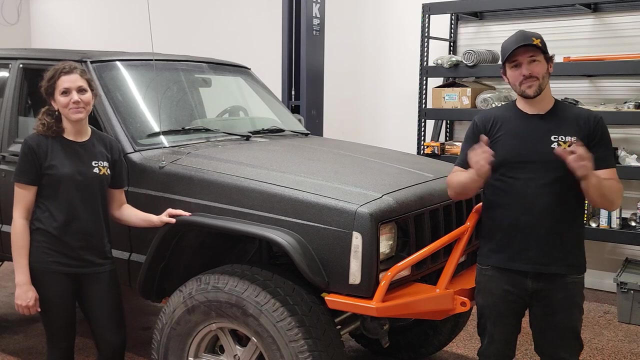 Spence and Marissa standing with the Rhino-lined XJ Cherokee in the Core 4x4 shop, orange bumpers visible
