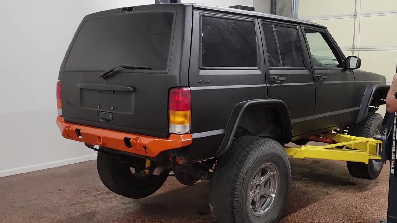 Rear view of the XJ Cherokee on the lift showing the Rhino line finish, orange Dirt Bound rear bumper, and Bushwacker flat flares