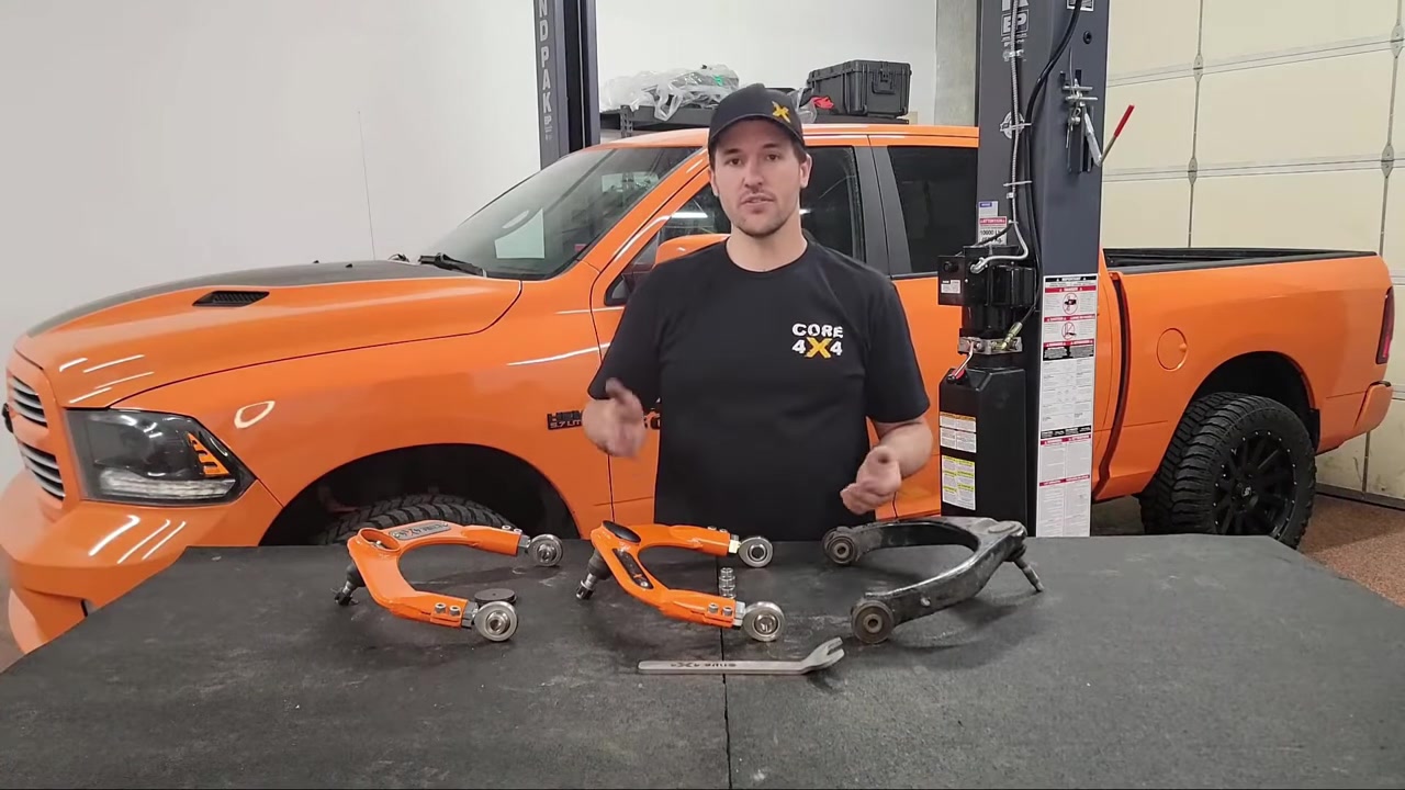 Spence showing the Core 4x4 adjustable upper control arms next to the factory arms on the workbench in front of the orange Ram 1500