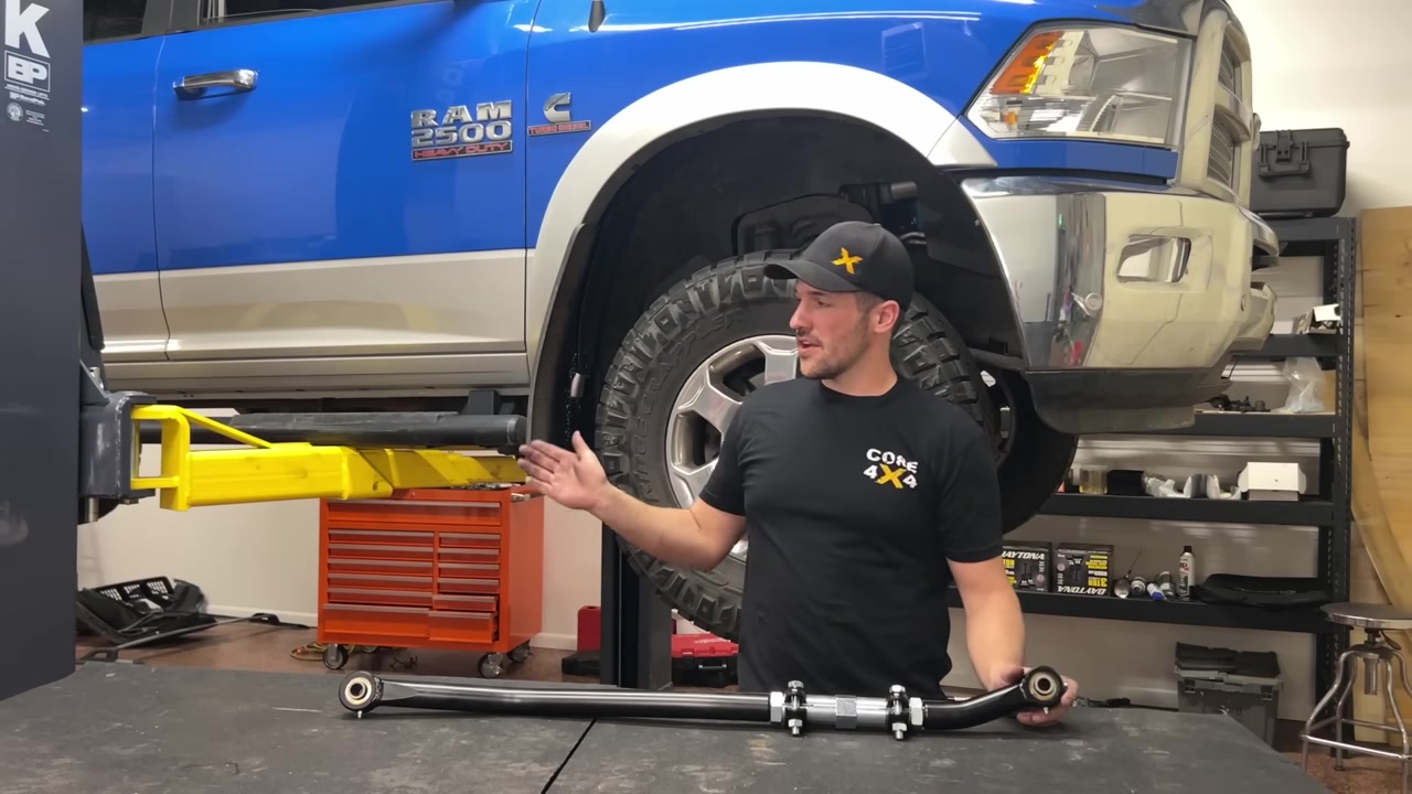 Spence holding the Core 4x4 Tow Series adjustable track bar next to the blue Ram 2500 on the lift, showing the turnbuckle adjuster and Johnny Joints