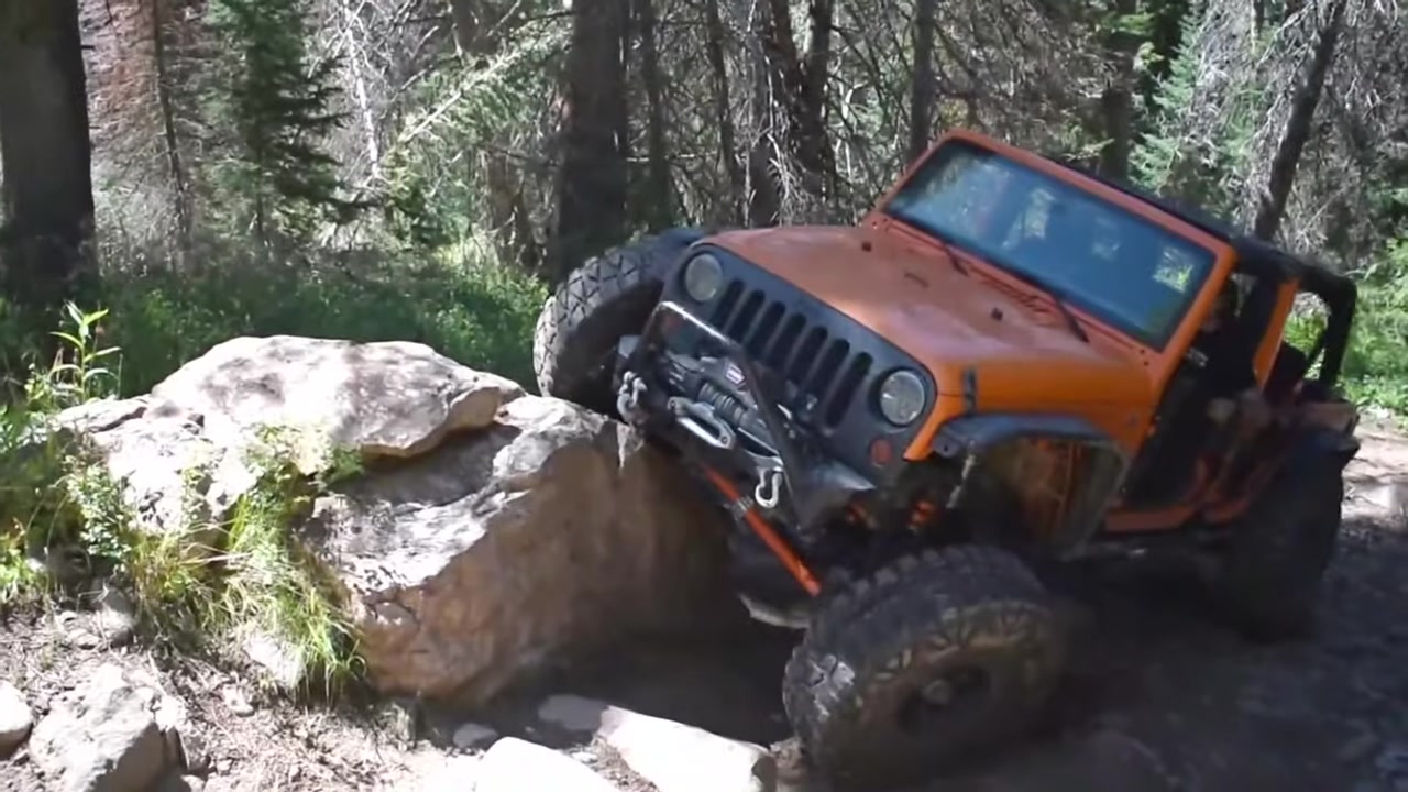 Orange Jeep JK with Core 4x4 suspension components crawling over large rocks on a forest trail, front suspension fully compressed showing the need for proper bump stops