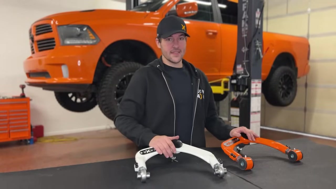 Spencer from Core 4x4 comparing the factory white upper control arm to the orange Core 4x4 adjustable arm on the workbench with the orange Ram 1500 on the lift behind him