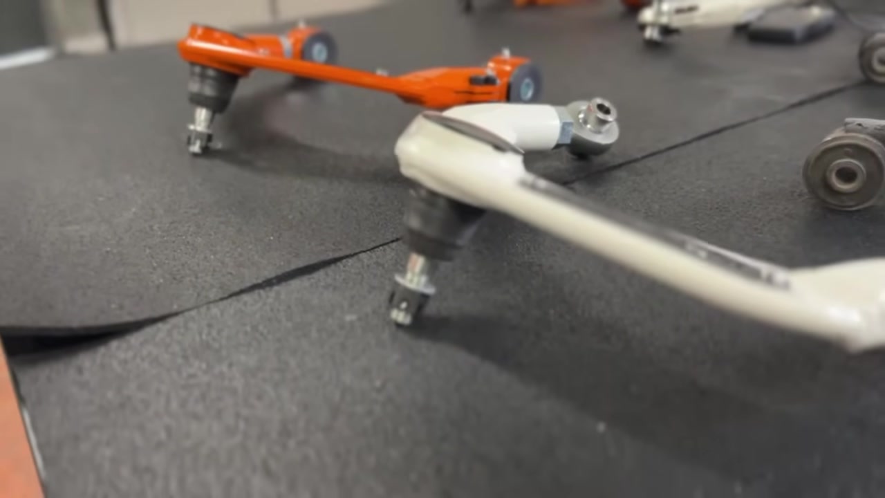 Close-up of the Core 4x4 adjustable upper control arm on the workbench showing the ball joint, adjustment sleeve, and offset weld angle alongside the orange arm