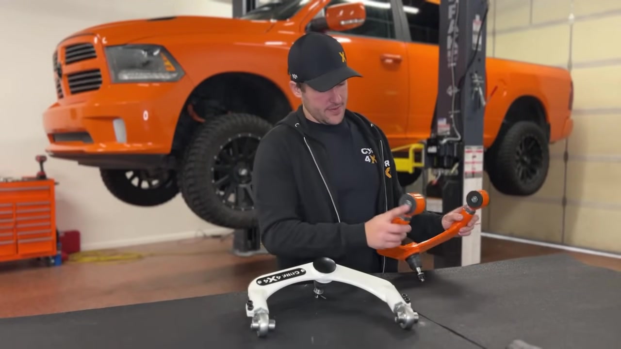 Spencer showing the FK rod end upgrade option on the Core 4x4 upper control arm, holding the white factory arm with the orange Core 4x4 arm on the workbench