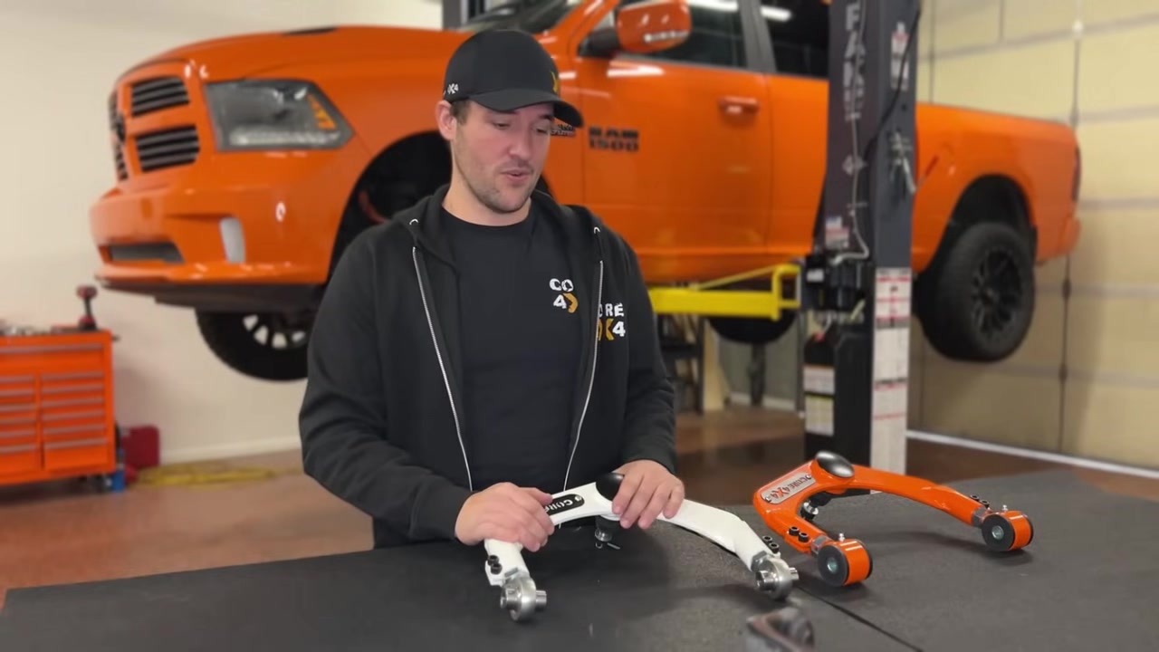 Spencer from Core 4x4 in the studio with the factory and Core 4x4 adjustable upper control arms on the workbench, orange Ram 1500 on the lift behind him