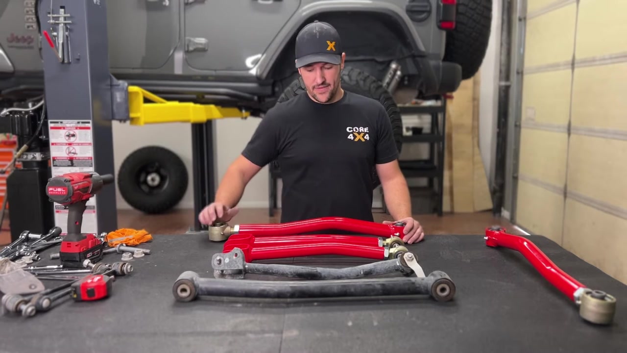 Spence measuring Core 4x4 Crawl Series control arms on a workbench with red arms and factory arms side by side