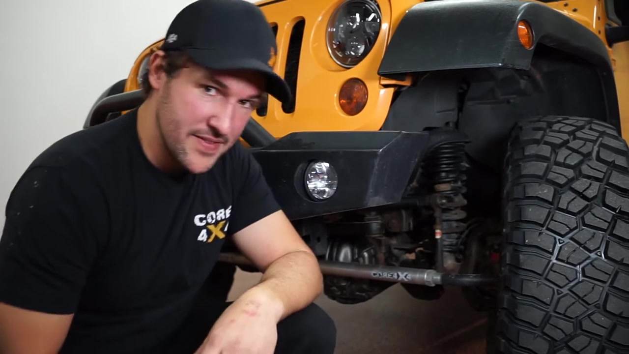 Spence kneeling next to the orange JK Wrangler pointing to the front sway bar and factory end link location on the driver side