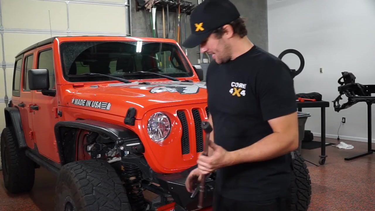 Spence demonstrating the passenger side bracket spreading technique using a breaker bar on a red JL Wrangler factory axle in the Core 4x4 shop