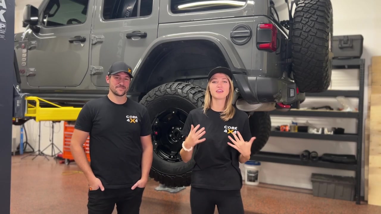 Gabby and Spence from Core 4x4 standing in front of the gray JL Wrangler on the shop lift before the rear control arm and track bar install