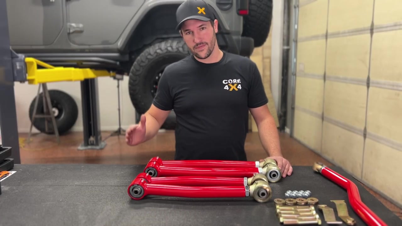 Spence at the workbench with the full Core 4x4 Crawl Series rear control arm kit laid out in red powder coat, showing the upper and lower arms, track bar, Johnny Joints, and hardware kit for the JL Wrangler