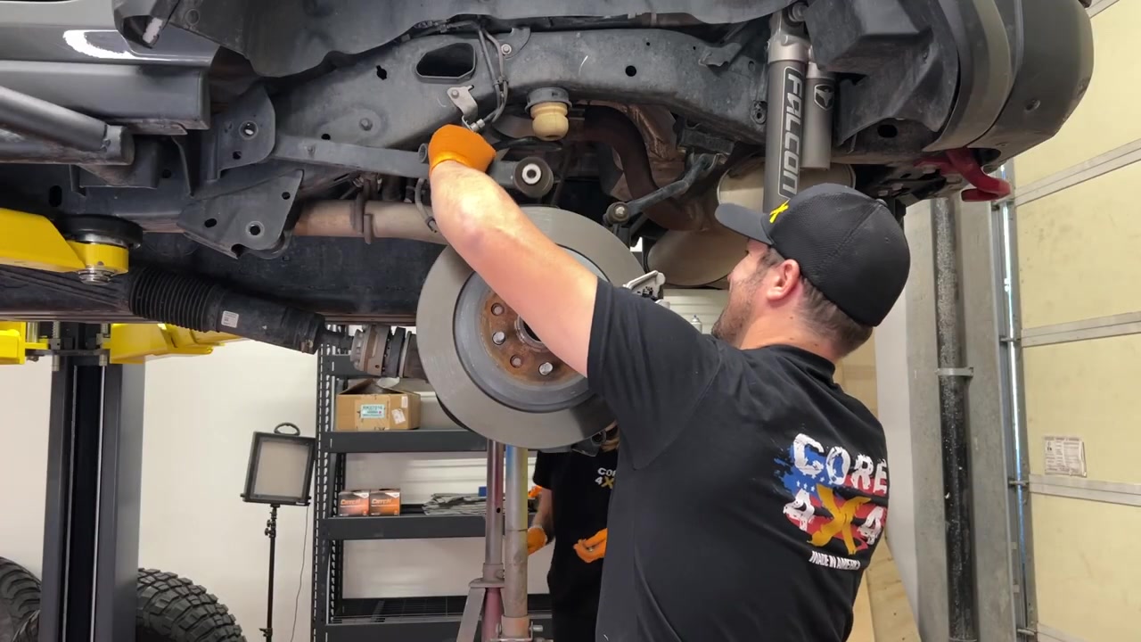 Spence under the JL Wrangler on the lift disconnecting the rear suspension components, with the Falcon shock and rear axle visible