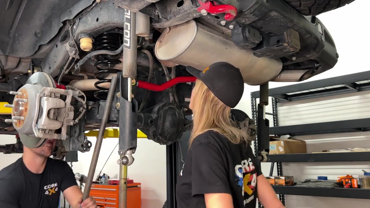 Gabby and Spence under the JL Wrangler inspecting the newly installed red Crawl Series rear control arms, taller springs, and track bar on the rear axle