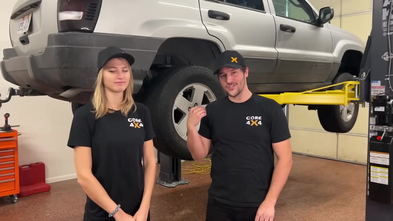 Spence and Gabby from Core 4x4 standing in front of the silver Jeep Grand Cherokee WJ on the lift before the rear upper A arm install