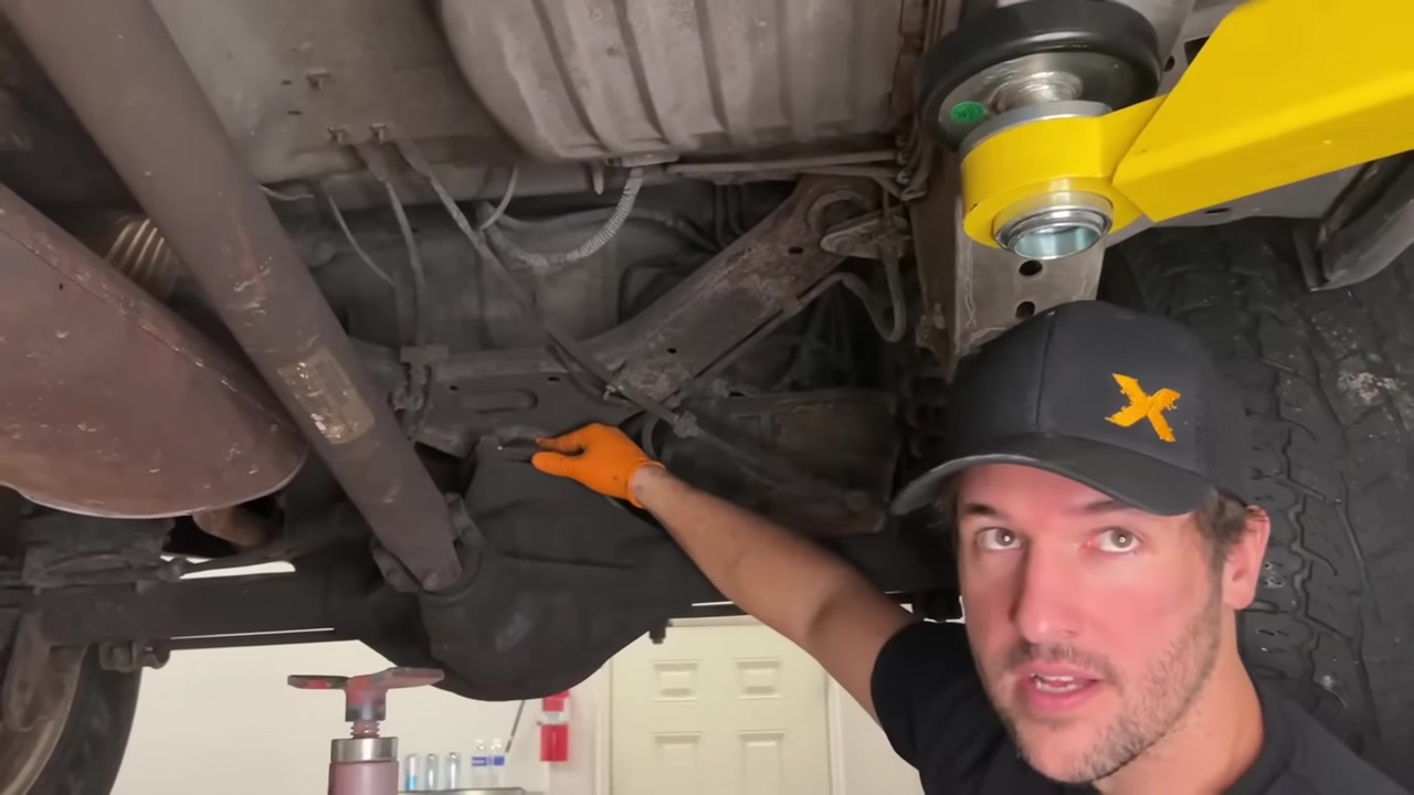 Spence under the WJ pointing at the factory rear upper A arm during removal, showing the three bracket bolts and brake line routing