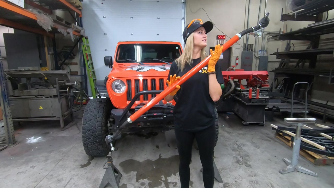 Gabby holding the Core 4x4 orange 7075 aluminum tie rod in front of the JL Jeep Wrangler