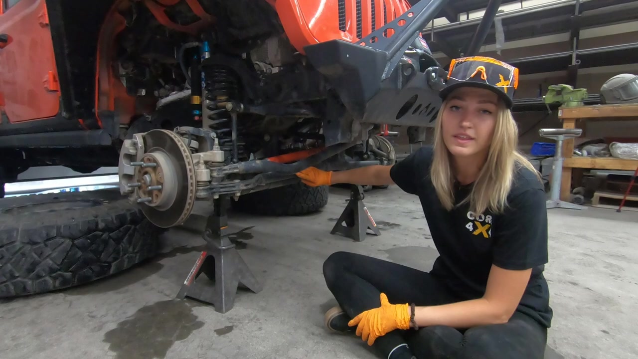 Gabby sitting next to the JL Wrangler on jack stands with the front axle and OEM steering visible
